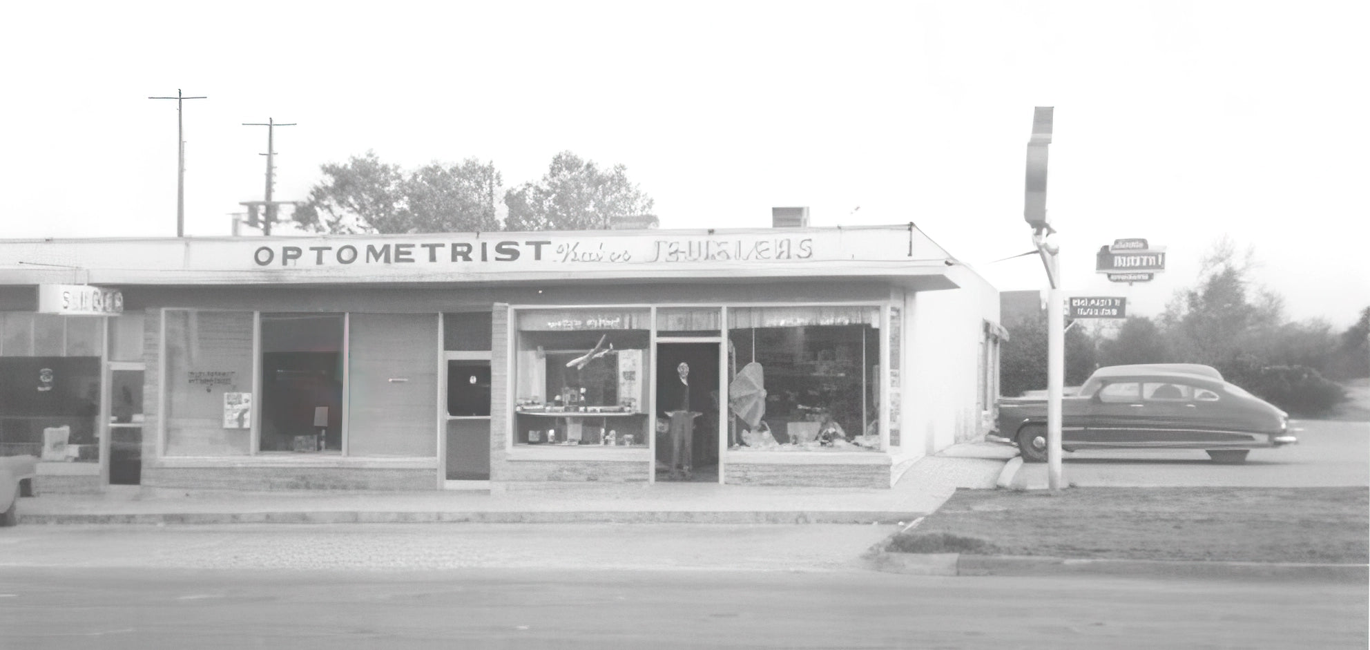 Vintage black and white photo of an optometrist office and Kubes store with a car parked outside.